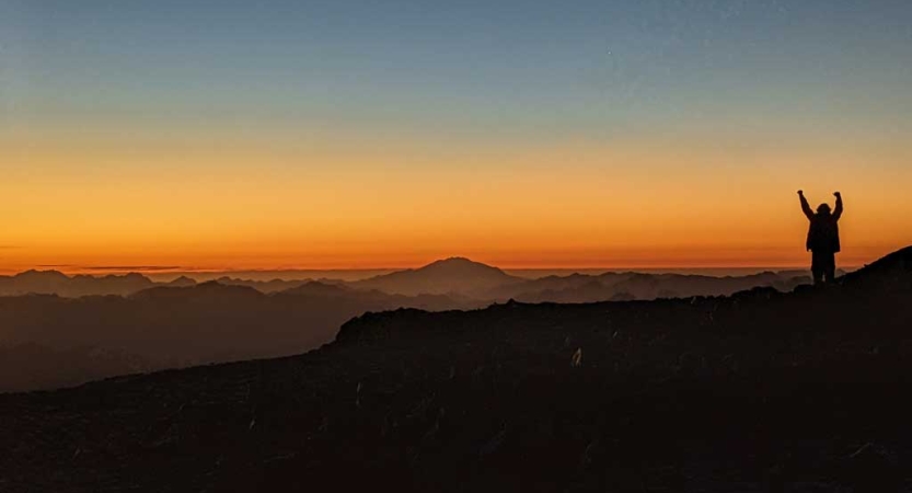 A person stands on a mountainous overlook and raises their hands into the air. The sky appears in shades or orange, yellow and blue, as the sun is either rising or setting. 